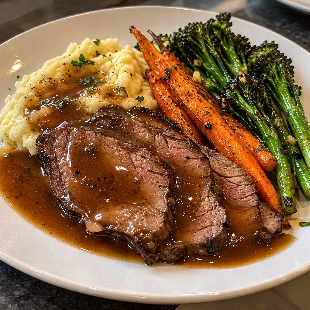Sliced roast beef with gravy, mashed potatoes, roasted carrots and broccolini on a white plate